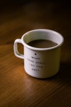white and black ceramic cup with 'what good shall I do to this day' on it - filled with brown liquid on brown wooden surface