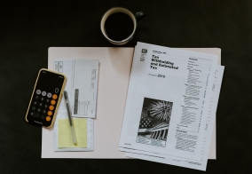 checkbook duplicate, post-it note, and white printer papers showing text about taxes in a beige folder on a black surface with a cup of coffee and iPhone in a yellow case with calculator feature